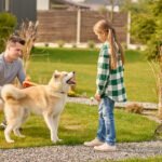 Acquaintance. Smiling middle-aged man in casual clothes crouching near dog with leash and school-age girl standing sideways to camera looking with interest fine afternoon